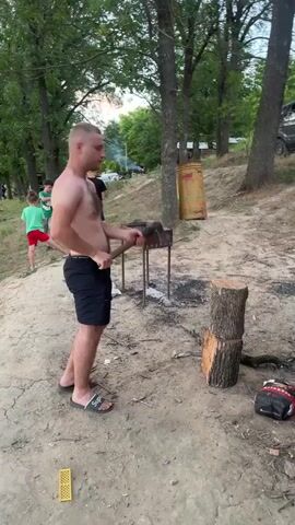 A shirtless man in black shorts and flip-flops is chopping a log with an axe. He is standing on a dirt path next to a barbecue grill. There are trees in the background, and a few children are standing to the left of the frame watching. The man swings the axe and then brings it down on the log.
