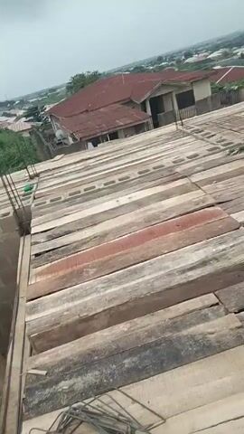 The video depicts a construction site, apparently on the roof of a building under construction. The roof is covered in wooden planks, with reinforcing steel rebar cages laid out on top. A man wearing a green shirt is seen working on the structure. The surrounding area shows a dense residential neighborhood with various buildings, trees, and an overcast sky. The camera moves, providing different perspectives of the construction site and the surrounding cityscape.
