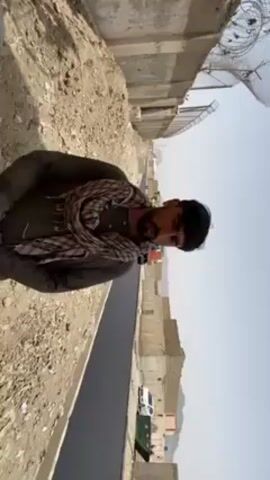 The vertical video captures an outdoor scene, likely a road construction or development site in an arid region.

In the foreground, a man with a dark beard and mustache looks directly at the camera. He wears a dark olive-green long-sleeved shirt, possibly a local garment like a *shalwar kameez*, and a patterned light-colored scarf (resembling a *shemagh* or *kufiya*) wrapped around his neck. His expression is neutral.

Behind him, a newly paved, dark asphalt road stretches into the distance, bordered by a white concrete curb on the left. Further down the road on the left, another man in a dark top and light cap is visible, bent over and appearing to be working on the road. A white pickup truck is parked much further down the road on the right.

The surrounding landscape is barren and dusty, with rocky, uneven ground and scattered debris lining the sides of the new road. Concrete walls or barriers run alongside parts of the road. In the mid-distance on the right, several simple, unfinished, or somewhat dilapidated buildings are visible. A large, white parabolic antenna or satellite dish is perched atop a structure on the right side of the frame.

The sky is clear and pale blue, indicating bright daylight, possibly midday or early afternoon, with strong shadows suggesting intense sunlight. The overall atmosphere is one of ongoing infrastructure work in a rugged, desert-like environment, consistent with Kandahar, Afghanistan. No discernible text is visible in the frame.