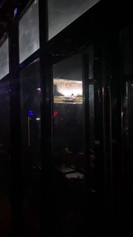 The video shows a television screen through dark rectangular frames. On the screen are several images of women in different poses and outfits, including a woman standing with a hula hoop and a woman sitting on a chair. The TV is in a dark bar or club-like setting. There is a red neon sign at the bottom of the screen.