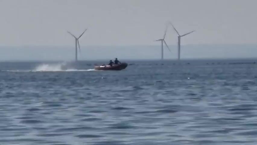 The video shows a view of the sea with a small inflatable boat carrying two people. The boat is moving from left to right, leaving a wake behind it. In the background, there is a distant coastline with several wind turbines. The sea is choppy with moderate waves. The sky is overcast, suggesting a possibly hazy or cloudy day. The overall scene appears to be a quiet, open-water environment, possibly offshore. The presence of wind turbines suggests an area where renewable energy generation is significant. The activity is simple transit by boat. No text is visible in the video.