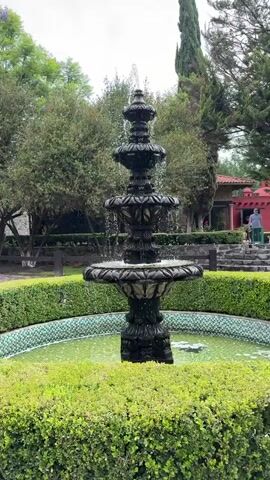 A full shot shows a three-tiered fountain in a garden. The fountain is black with intricate designs, and water sprays from the top tier and cascades down each level. The water in the fountain's basin is a cloudy green. A short, trimmed hedge surrounds the fountain, forming a semicircle at the bottom of the frame. In the background, green trees and foliage are visible. To the right, a person in a blue shirt stands near a red building with a red roof, and stone steps lead up to the building. The sky is overcast.