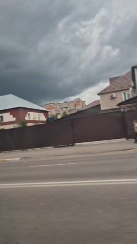 The video shows a car driving past buildings and a street. The sky is cloudy. In the first clip, a two-story building with a large glass entrance is visible. In the second clip, several cars are parked along the street in front of brick buildings with red roofs. One building has a blue roof. In the third clip, a dark-colored SUV drives past a pink two-story house. In the fourth clip, a silver sedan drives past the pink house, and a person walks on the sidewalk. In the fifth clip, a person walks on the sidewalk near a brick building.