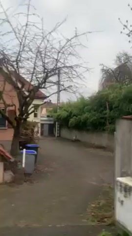 The camera is panning along a row of houses, with a fence in the foreground. The houses are mostly white with dark-colored window frames and roofs, and some have small yards with greenery. The sky is overcast.