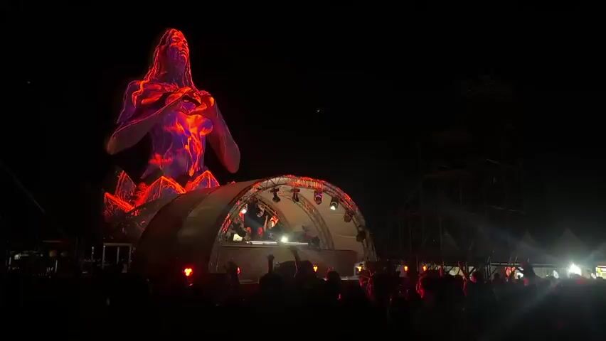A nighttime outdoor music festival is in progress. A large, stylized statue of a human figure is illuminated with projected patterns and colors. In front of the statue, a stage with a curved roof is set up, featuring lighting equipment and a DJ booth. A crowd of people is gathered in front of the stage, silhouetted against the stage lights and the projections on the statue. Some members of the crowd are raising their hands. Red lights and beams are visible, pulsing with the music. The statue's projection changes throughout the clip, transitioning from white and floral patterns to fiery orange and then back to a translucent, icy appearance. The overall atmosphere is energetic and celebratory.