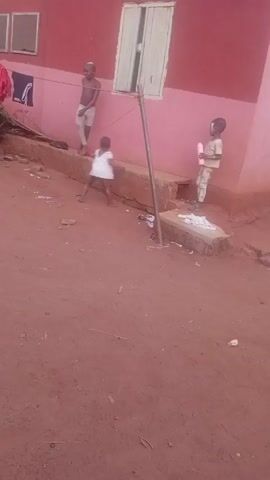 The video shows three children playing outside a pink building with white window frames. The ground is red dirt. One child is shirtless, wearing light-colored shorts, and dribbling a basketball. Another child, wearing a white shirt, runs in the same direction. The third child stands near the building holding a cylindrical object, possibly a bottle.  There is a metal pole standing upright, as well as litter scattered on the ground. Clothes are hanging on a line in the background.
