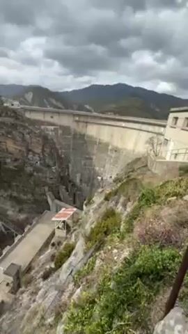 The video shows a person rappelling down the side of a cliff near a large dam. The dam is made of concrete and is situated in a mountainous area. The person rappelling is wearing a helmet and dark clothing and is attached to a system of ropes and cables. The camera is positioned above the person, looking down at them as they descend. The weather appears to be overcast. At the base of the cliff, there are some buildings and a small pool of water.