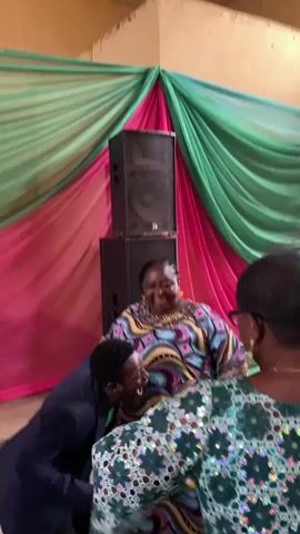 This is a video of two people dancing at an event. A man in a blue suit is on the left, leaning towards a woman in a colorful dress who is laughing and dancing. In the background, there are stacked speakers and green and pink draped fabric. A third person, partially visible on the right, appears to be wearing a green and silver patterned outfit. The lighting suggests an indoor setting, possibly a hall or event space. The overall atmosphere is joyful and celebratory.