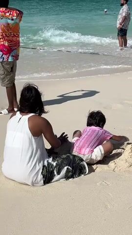 Two people are sitting on a sandy beach near the ocean. A woman in a white dress and a child in a pink shirt and white shorts are playing in the sand. A man in a brightly colored shirt and khaki shorts is standing behind them, taking a photo with his phone. Further out in the ocean, another man in a patterned shirt and shorts is standing in the water. The water is turquoise and waves are gently rolling onto the shore. The sky is bright and sunny, casting shadows on the sand.