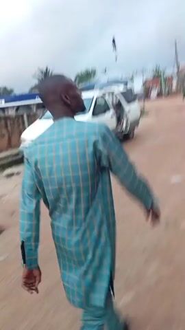 This is a short video captured in Idimu, Nigeria, appearing to be a roadside scene with a gas station and a dirt road. The weather seems overcast, suggesting it might be morning or a cloudy day.

The primary subjects are people and vehicles. A white minivan is parked near what appears to be a Mobil gas station. In the foreground, a person wearing a light blue and green striped traditional African garment is walking away from the camera. The person's movement is slightly blurred, indicating motion. Further in the background, other vehicles and buildings are visible, including what looks like more gas pumps and storefronts.

The activity captured is the movement of people and vehicles in a public area. The person in the foreground is walking along the dirt road, seemingly heading towards or away from the gas station area. The scene evokes a sense of everyday life in a Nigerian town.

Notable details include the presence of the Mobil branding on the gas station, suggesting it's a recognizable chain. The dirt road and the style of the buildings point to a typical urban or semi-urban environment in Nigeria. The overcast sky contributes to a muted color palette of the video. There is no discernible text that is clear enough to read.