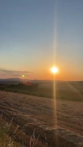 The video shows a wide shot of a sunset over a rural landscape. The sun is a bright, radiant orb low in the sky, casting a warm glow over the scene. The sky transitions from a pale blue at the top to a soft orange and yellow near the horizon. In the foreground, a field of dry, golden grass is visible, with the stalks bent and intertwined. Beyond the field, rolling hills recede into the distance, dotted with trees and small buildings. The overall impression is one of peacefulness and natural beauty. There are no people or animals visible in the video. The lighting suggests it is late afternoon or early evening.