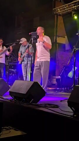 A man in a light colored shirt and gray pants stands center stage, singing into a microphone. He is bald and has his eyes closed. To his left, another man in a white shirt and blue jeans plays a guitar. To the far left, a man in a white shirt and sunglasses plays a bass guitar. Behind the singer, a keyboard is visible. The stage is lit with blue and green lights.
