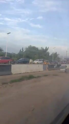 The video shows a view from inside a vehicle looking out a window at a street scene. The sky is cloudy and light blue. There are trees and utility poles visible in the background. On the street, there are several cars, including a red SUV and a yellow tricycle-like vehicle. Some people are standing near the vehicles and on the sidewalk. There is a concrete barrier separating the road from the area where the people are. The ground is dirt and gravel. The video appears to be taken in the daytime.