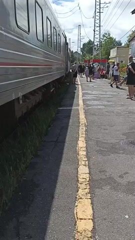 The video shows a stationary train on the left side of the frame, with its silver carriages visible. On the right side of the frame is a train platform, paved with asphalt and marked with a yellow line. Several people are standing and sitting on the platform. Some are holding luggage. In the background, there are buildings, trees, and utility poles with overhead wires. The sky is partly cloudy. The lighting suggests it is daytime.