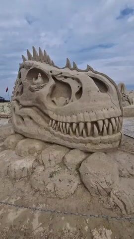 A slow pan shows a large sand sculpture of a dinosaur skull, possibly a Tyrannosaurus Rex, resting on a mound of sand. The sculpture is intricately detailed, with sharp teeth and eye sockets. The background is a cloudy sky with some trees and other sand sculptures visible in the distance. There are no people visible in the frame. The sculpture is surrounded by a chain fence.
