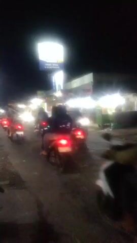 The video shows a night scene on a road filled with motorcycles. The motorcycles are moving, and their headlights and taillights are visible. Some of the motorcycles have two people riding them. There are also some buildings or stalls with bright lights in the background. The overall impression is a busy street at night.