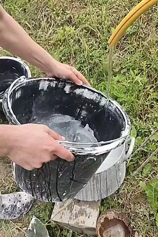 A person is shown pouring the contents of a black plastic bucket into a large metal pot. The black bucket appears to have a white residue inside, and the person is holding it with both hands. The metal pot is already partially filled with a white, frothy liquid, and a yellow garden hose is visible at the top right of the frame, with water flowing from it into the pot. The scene appears to be outdoors, with grass and vegetation in the background. There are also other objects visible in the foreground, including a pair of white and grey patterned clogs and another black bucket on the left.