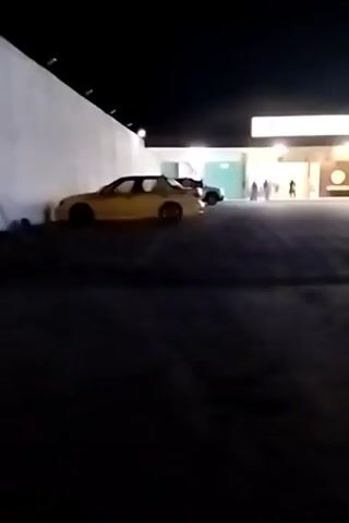 A person is walking in an outdoor area at night. The person is walking past a yellow car that is parked near a white wall. The person continues walking towards a building with bright lights and other people visible. There are also other cars and motorcycles parked in the area. A sign with text in what appears to be an Asian language is visible on the building.