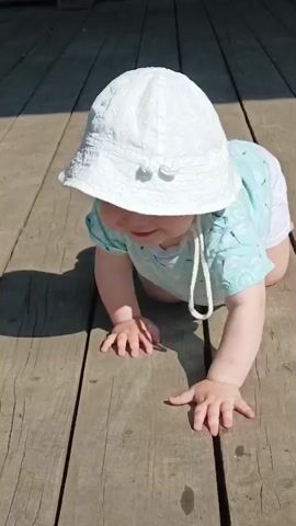 A baby wearing a white sun hat and a light blue t-shirt with a dragonfly pattern is crawling on a wooden deck outdoors. The baby is on its hands and knees, facing forward, with its head turned slightly to the side. Sunlight casts a shadow of the baby on the deck. In the background, a green wooden wall is visible. The video is shot from a low angle, close to the baby.