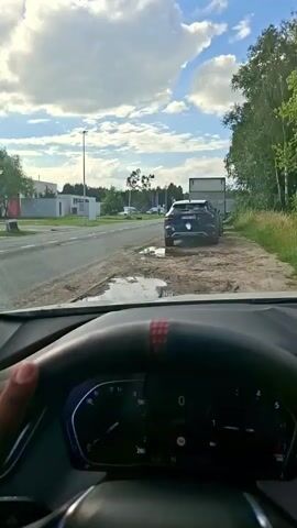This video shows the interior of a car, looking out through the windshield. The camera is positioned behind the steering wheel, which has a BMW logo in the center and a red and black stripe near the top. The dashboard displays speed and other vehicle information.

Outside the car, it appears to be daytime with a partly cloudy sky. The scene is a roadside with a paved road on the left and a dirt shoulder on the right. There are some buildings in the distance on the left, and trees and vegetation on the right. A dark-colored SUV is parked on the dirt shoulder in front of a large white shipping container. There are puddles of water on the ground near the parked SUV.

The activity taking place is driving, as indicated by the view from the car and the dashboard. The car appears to be moving slowly. The overall mood is neutral and observational. There is no discernible text visible in the video.
