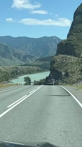 The video shows a view from a vehicle traveling on a paved road. The road curves to the right and descends slightly. To the left of the road is a turquoise river flowing through a valley. The valley is surrounded by lush green mountains. On the right side of the road, a steep, rocky cliff face rises sharply. A dark-colored SUV is in front of the camera vehicle, also traveling on the road. The sky is blue with a few scattered white clouds. There are signs on the right side of the road, including one with a circular shape and the number 50, indicating a speed limit. The overall impression is a scenic drive through a mountainous region with a river.