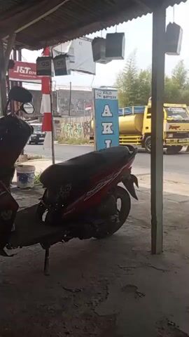 The video shows a street scene from a covered area, possibly a shop or a shelter. In the foreground, a red and white motorcycle is parked. To the right of the motorcycle, there is a wooden structure and part of a woven seat. In the background, across a paved road, there are several buildings and vehicles. A large yellow tanker truck with a blue tarp covering its tank is moving to the right. There are also some smaller white trucks and a pink vehicle that appears to be a bus. Signs are visible on some of the buildings, including one with the text "FZ BATTERY" and another with text that includes "SALON". Graffiti is present on a wall to the left of the road. The sky is bright, suggesting it is daytime. The camera is stationary.