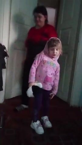 A young girl stands in a doorway wearing a pink Peppa Pig hoodie, purple leggings, and white sneakers. She is holding a black and white stuffed animal. A woman stands behind her, partially obscured. The girl looks around and gestures with her hands. The doorway leads to another room. The walls are painted white. A coat hangs on a coat rack to the left of the doorway. The floor is dark.