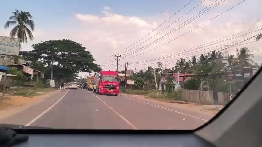 The video shows traffic on a two-lane road in what appears to be a rural or semi-rural area. The view is from inside a car, looking forward through the windshield. A large red truck with a tank is prominent in the initial frames, followed by a yellow truck. Other cars and motorcycles are also visible on the road, moving in both directions. There are trees, utility poles, and houses lining the side of the road, with a mix of cloudy and clear sky overhead.