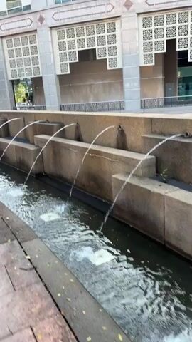 The video pans from left to right along a decorative fountain. Water flows from multiple spouts, creating arcs that fall into a channel. The fountain is built with tiered concrete structures, and behind it is a tan-colored building with ornate, patterned window coverings. Trees and other buildings are visible in the background. The foreground shows a paved walkway with a red track running alongside it. The lighting suggests it is daytime. There are no people visible in the video.