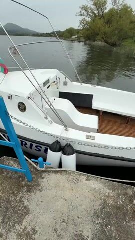 The camera pans upward along the side of a white boat docked beside a gray concrete pier with a blue railing. Two white cylindrical boat fenders are visible, secured to the boat with rope and chains. The boat has wooden flooring in the stern. A Bulgarian flag is affixed near the bow of the boat. A tall white light post stands on the pier beside the boat. In the background, there is a calm lake and a treeline, with mountains visible in the distance. The sky is overcast and gray.