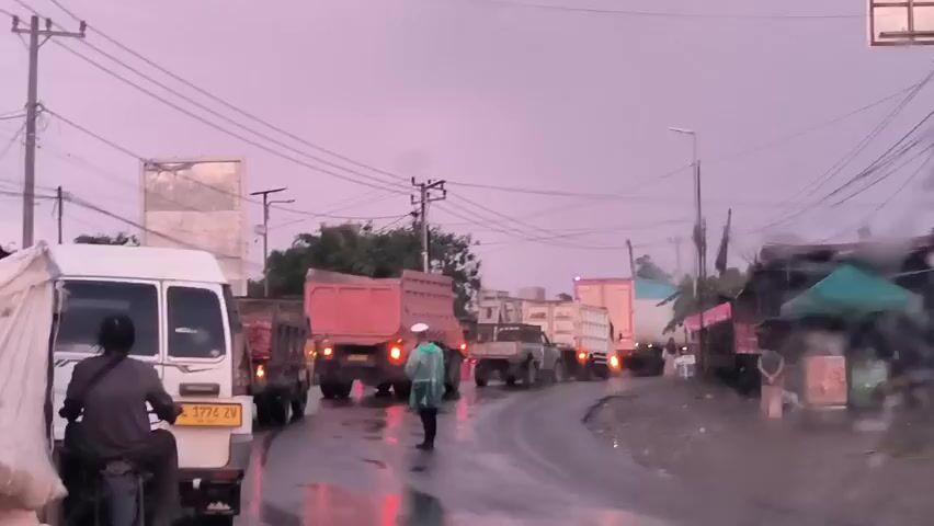 The video shows a traffic jam on a wet road, likely after a rainfall. A man wearing a light green raincoat is standing in the middle of the road, apparently directing traffic. Several trucks loaded with goods are lined up ahead, their brake lights illuminated. A white van and a motorcyclist are visible in the foreground on the left side of the road. The sky is overcast, suggesting it's either early morning or late afternoon. Power lines crisscross overhead. A few small shops or stalls are on the right side of the road, some with green awnings. The overall scene suggests a busy but somewhat disorganized environment.