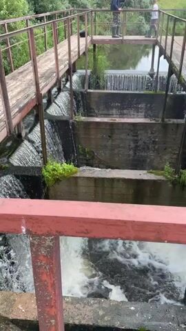 The video shows a tiered water structure, possibly a dam or a series of sluice gates, with water cascading down several levels. Two people are visible on a walkway above the structure, one appearing to be fishing. The structure is made of concrete and metal, with wooden planks forming the walkway. Lush green vegetation surrounds the water. The overall environment appears to be rural, with trees and hills in the background. The water is choppy and white, indicating a significant flow. The perspective is from a slightly elevated position, looking down onto the water structure and the people on the walkway.