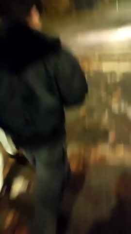 The video shows two people walking down a sidewalk at night, then the camera pans to a street with a crosswalk. The street is wet and there are leaves on the ground. There are streetlights and traffic lights visible, and cars are driving on the road. The video is shaky and the focus is not always clear.