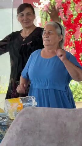 Two women are captured in a medium shot, facing each other with a backdrop of vibrant pink and red flowers. The woman on the left wears a dark, patterned dress and has dark hair pulled back. She is smiling and gesturing with her left hand. The woman on the right is older, with long grey hair braided down her back, and she is wearing a blue dress. She is looking away from the camera, her right hand raised to her head. A third person, partially visible on the right edge of the frame, appears to be clapping. The foreground shows a blurred section of a light-colored, textured surface, possibly a piece of furniture. The setting seems to be an indoor event or celebration, indicated by the decorative floral arrangement.