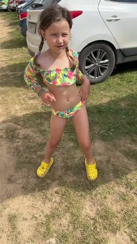 A young girl with braided brown hair is standing and dancing outdoors. She is wearing a floral print two-piece swimsuit with long sleeves and yellow Crocs. The girl is positioned on a patch of grass with a dirt path and parked cars in the background. The sun is shining.