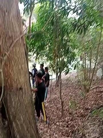 A group of five young males are gathered near a body of water in a wooded area. The ground is covered with fallen leaves. One male in the foreground is wearing a black t-shirt and yellow-striped pants. Another male is wearing a black shirt and hat. The others are also wearing dark clothing. The environment appears to be a jungle or a similar dense, natural setting.