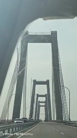 The video is shot from the interior of a car driving across a cable-stayed bridge. The bridge has tall, concrete towers connected by numerous cables, and the road is divided by lines with several cars moving in the same direction. Streetlights line the sides of the bridge. The sky is overcast and bright. The corner of the car's windshield and part of the dashboard are visible in the foreground. The watermark "Infinix NOTE 40 Pro" appears at the bottom of the video.