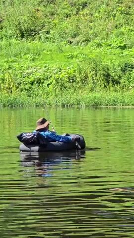 A person in a brown hat and blue shirt is fishing from a black inflatable tube in a body of water. They are holding a fishing rod. Another person is swimming in the water in front of the inflatable tube. The background is a lush green hill.