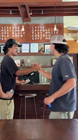 The video shows a small coffee shop. Two customers are at the counter, one looking at something on his phone. Another customer in a red shirt approaches the counter. Behind the counter stands an employee with a baseball cap. A menu is mounted to the wall, listing different coffee drinks. Various items are on display on the shelf behind the counter including mugs, artwork, and a small Marshall speaker.