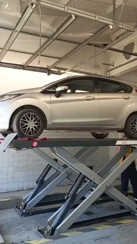 A silver Ford Fiesta is slowly being lowered on a hydraulic car lift. A person in a dark blue uniform and a yellow safety vest is standing to the right of the lift, observing the process. The car lift is located inside a building with a metal roll-up door overhead and bright lighting. The scene appears to be a car maintenance or inspection area.