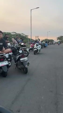 The video captures a scene on a street in Besuki, Indonesia, likely during sunset due to the warm, hazy sky. A group of people are gathered on the side of the road, many sitting on or near parked motorcycles. The motorcycles range from classic designs to modern scooters, with one prominent white scooter in the foreground. Some individuals appear to be socializing or waiting, leaning on a low wall or railing that separates the road from a vegetated area.

Further down the road, more people and motorcycles are visible, suggesting a communal gathering or a popular spot for people to congregate. The road itself appears to be a main thoroughfare, with traffic continuing in the distance. The overall atmosphere seems relaxed and casual, with the golden light of dusk casting a soft glow over the scene. The time of day is evident from the low angle of the sun and the muted colors in the sky. There are no visible text elements within the video.