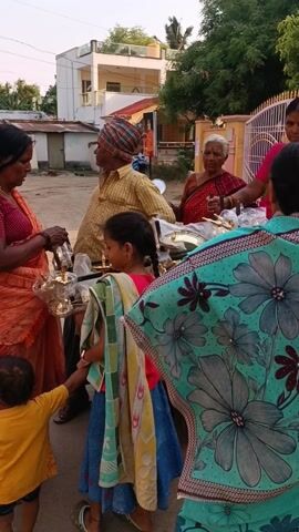 Here is a description of the video:

The video shows a street scene with several people gathered around a cart selling small, brass oil lamps. The vendor, an older man wearing a turban and a yellow striped shirt, stands next to the cart. Several women in saris are examining the lamps, including one wearing an orange sari, who is holding one of the lamps and talking to the vendor. A young girl in a red shirt and a blue skirt, with a towel draped over her shoulder, is standing near the cart, while a toddler in a yellow shirt stands next to the woman in the orange sari. An older woman in a red sari stands near a metal gate in the background. Houses and greenery can be seen in the background. The lamps on the cart appear to be new and shiny. The interaction suggests a transaction, with the people discussing the items for sale.