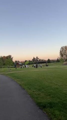 The video pans across a grassy park during sunset. In the distance, a group of people is playing volleyball on a court with a net. Further to the right, there is a playground with slides and other equipment. Several trees are scattered throughout the park, and a paved path curves into the foreground on the left. The sky is a clear blue with hints of orange and pink from the setting sun.