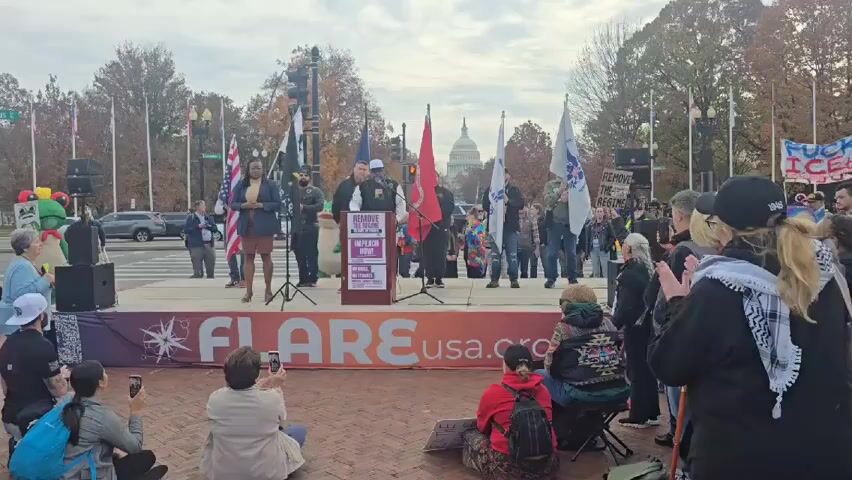 A rally is taking place in front of the US Capitol Building in Washington, D.C. A group of people are standing on a stage with a sign that reads "FLARE usa.org". There are several people speaking from the podium. A crowd of people are standing and sitting in front of the stage, some of whom are holding signs. Many are taking photos with their phones. The sky is overcast. There are several flags and banners displayed, including one that reads "F*CK ICE". There is a person dressed as a green character in the background. The location appears to be a public square, with streets and sidewalks visible. Cars and traffic are present.