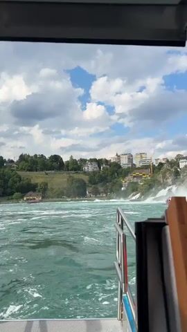 The video captures a scenic view from a boat navigating turbulent, turquoise waters. In the foreground, a silver railing and part of the boat's deck are visible. The mid-ground is dominated by the churning river, with white foam indicating its rapid flow. In the background, a lush green hillside is dotted with buildings and trees, leading up to a town or city. A prominent feature is a waterfall, with white water cascading down rocks. A pink boat with several passengers is positioned near the base of the waterfall, suggesting a tourist activity. Several people can be seen on a platform overlooking the falls. The sky above is a mix of blue and white clouds, indicating a partly cloudy day. The overall atmosphere is one of natural beauty and adventure, likely a daytime excursion at the Rhine Falls in Nohl, Switzerland. There is no visible text in the media file.