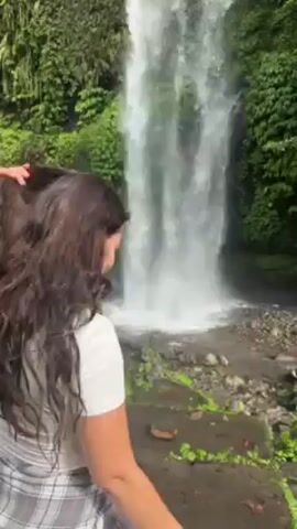 Two young women stand together in front of a waterfall. The waterfall is surrounded by lush green vegetation. One woman is wearing a white shirt and a gray and white plaid shirt tied around her waist. The other is wearing a pink and white striped shirt, a denim skirt, and a pink baseball cap. They are holding hands, smiling, and posing for the camera. The woman in the white shirt puts her hair up into a ponytail.