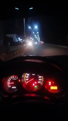 A car drives on a street at night. The car's dashboard is visible, and the speedometer reads approximately 40 km/h. Other vehicles can be seen on the road ahead, illuminated by streetlights. The car has a digital display showing music playing.