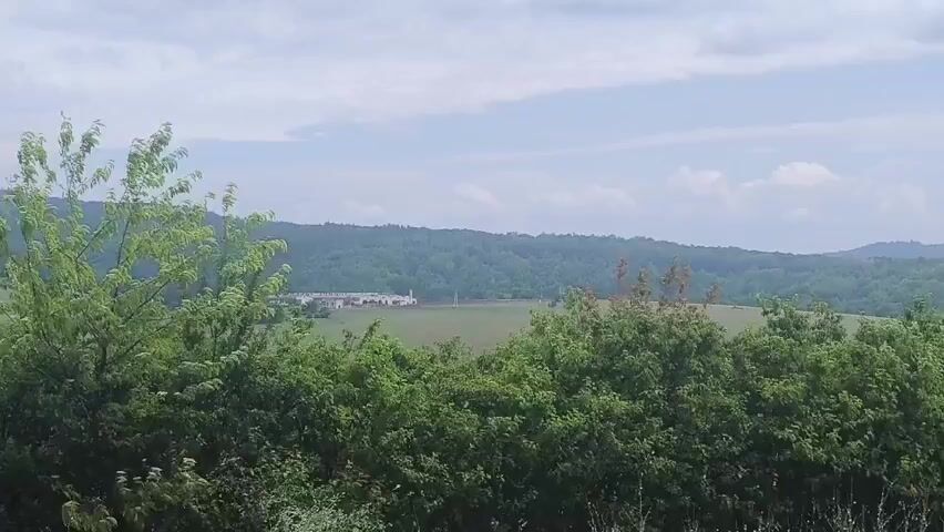 A wide shot of a rural landscape under a partly cloudy sky. In the foreground, lush green trees with some rust-colored leaves line the bottom of the frame. Beyond the trees, a wide, open field stretches towards a distant, treed hill. In the mid-ground, a long, white building complex with a central tower is visible in the field. The building appears to be some kind of institutional or industrial facility. The overall scene is peaceful and natural, with the man-made structure subtly integrated into the environment.