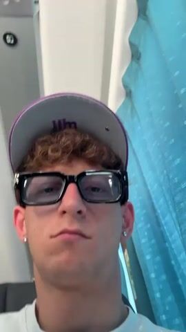 A young person with curly brown hair, wearing a purple and gray baseball cap, large black-rimmed glasses, and a white t-shirt, looks into the camera. The person has small hoop earrings in both ears and tattoos on their left hand. The background shows a blurred interior, possibly a bus or train, with blue curtains on the right and grey seating on the left. The person makes a peace sign with their left hand, briefly obscuring their face at 00:02, before resuming a neutral expression. The lighting suggests daytime.