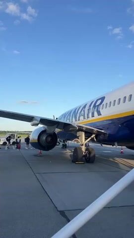 This video shows a Ryanair airplane parked on an airport tarmac. Passengers are visible in the foreground, some with luggage, and appear to be boarding or waiting to board the aircraft. The scene is set against a clear blue sky with some clouds, indicating it is daytime. The wing of another airplane is visible in the latter part of the video, as is an airport hangar in the background. The overall impression is one of an active airport environment during daylight hours.